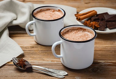 Dos chocolates calientes con leche junto a un plato con canela, anís y chocolate en tabletas