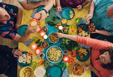 Vista cenital de comensales sirviéndose comida variada en mesa familiar y colorida.
