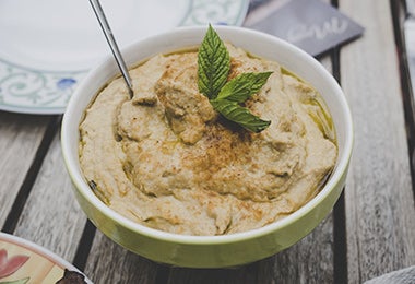 Hummus con unas hojas aromáticas y una cuchara en una mesa, un plato frío ideal de acompañamiento.