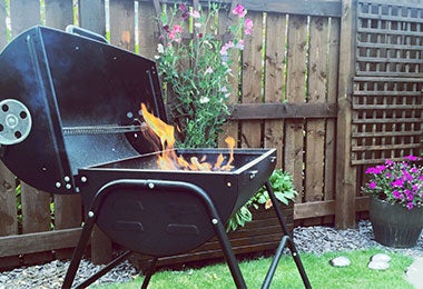 Parrillera metálica encendida en un jardín doméstico, rodeada de flores y una cerca de madera.
