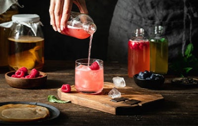 Vaso de kombucha rosada con hielo y frambuesa, servido desde una botella.