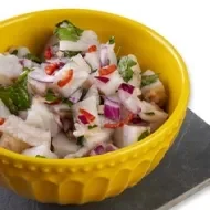 Ceviche con leche de tigre, cebolla morada, cilantro y limón en un recipiente amarillo sobre un fondo blanco