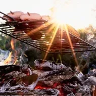 Parrilla metálica suspendida en fogón de leña, asándose sobre brasas ardientes.