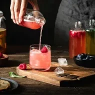 Vaso de kombucha rosada con hielo y frambuesa, servido desde una botella.