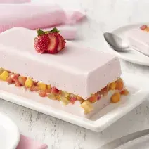 fondo blanco de madera con tela rosa claro, un plato blanco con una capa de gelatina con yogur de fresa, una capa de macedonia de frutas y otra capa de gelatina con yogur de fresa.