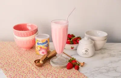 Fotografía de un batido rosa servido en un vaso alto. Al fondo, una escena con fresas y cuencos en tonos rosados.