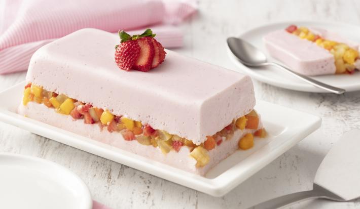 fondo blanco de madera con tela rosa claro, un plato blanco con una capa de gelatina con yogur de fresa, una capa de macedonia de frutas y otra capa de gelatina con yogur de fresa.